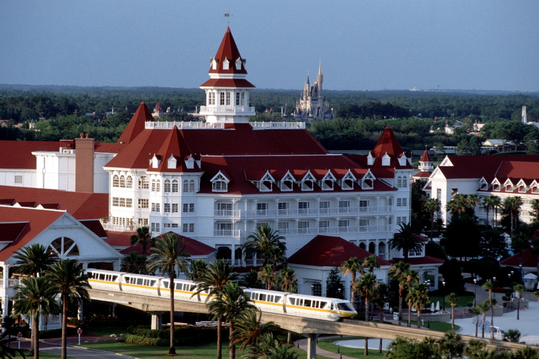 A train passing by a large white building with Disney's Grand Floridian Resort & Spa in the backgroundDescription automatically generated